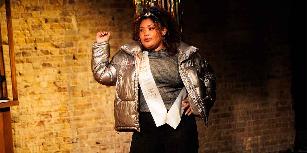 Olivia Forrest as Charlene poses confidently in a silver puffer jacket, with a sash reading "ALL ABOUT ME.