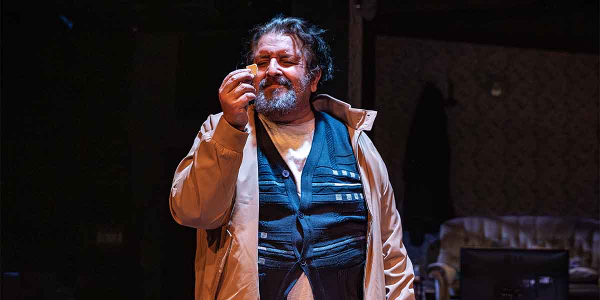 Rehan Sheikh as Liaquat holds a small object, smiling, in a dimly lit theatre setting.