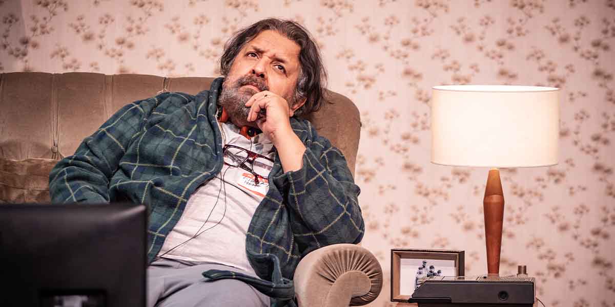 Rehan Sheikh as Liaquat sits thoughtfully on a sofa, wearing a green checked robe, with a lamp nearby.