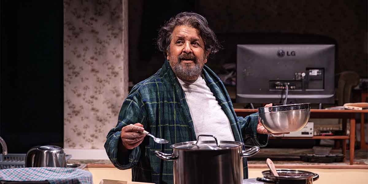 Rehan Sheikh as Liaquat, holding a bowl and spoon, stands in a kitchen setting with a pot.