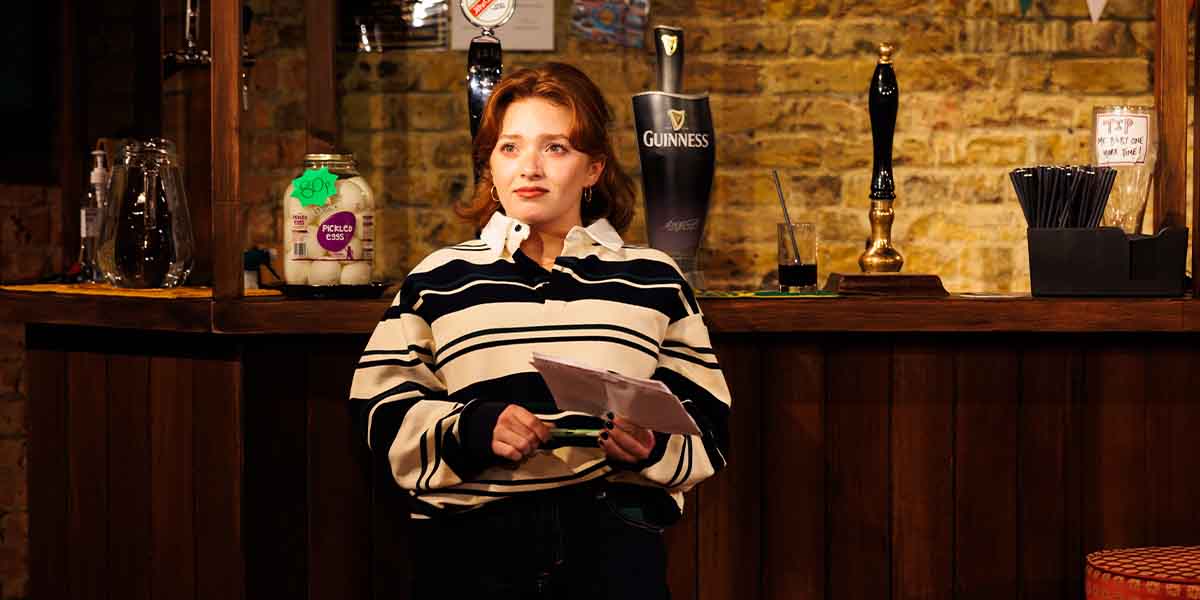 Rowan Robinson, wearing a striped shirt, stands at a bar holding a script, looking thoughtful.