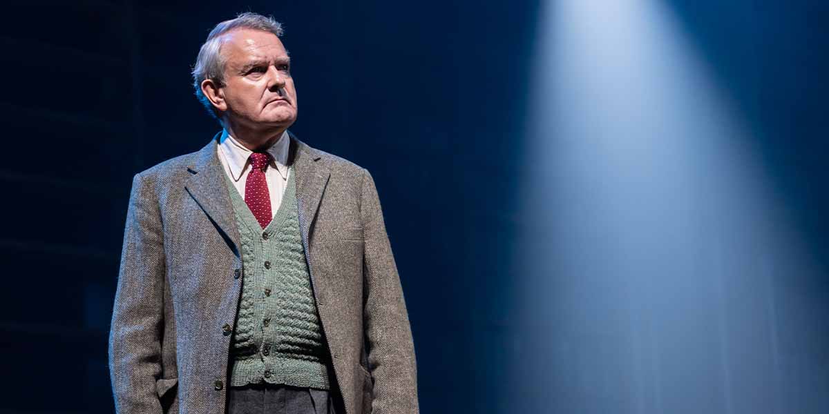 A man in a tweed jacket and green cardigan stands under a spotlight, looking serious.