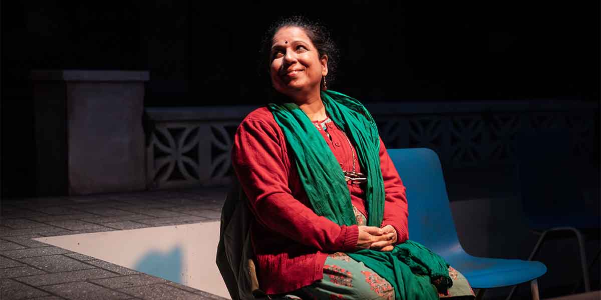 Shobu Kapoor as Hema smiles while seated, wearing a red top and green scarf, against a dark background.