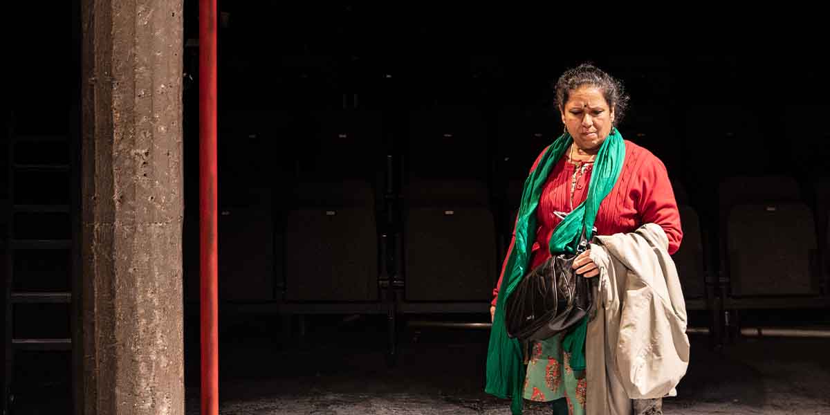 Shobu Kapoor as Hema, wearing a red top and green scarf, stands thoughtfully in a theatre.