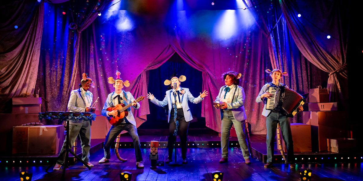 A ballet dancer in a pink tutu performs an arabesque on stage, with festive decorations in the background.