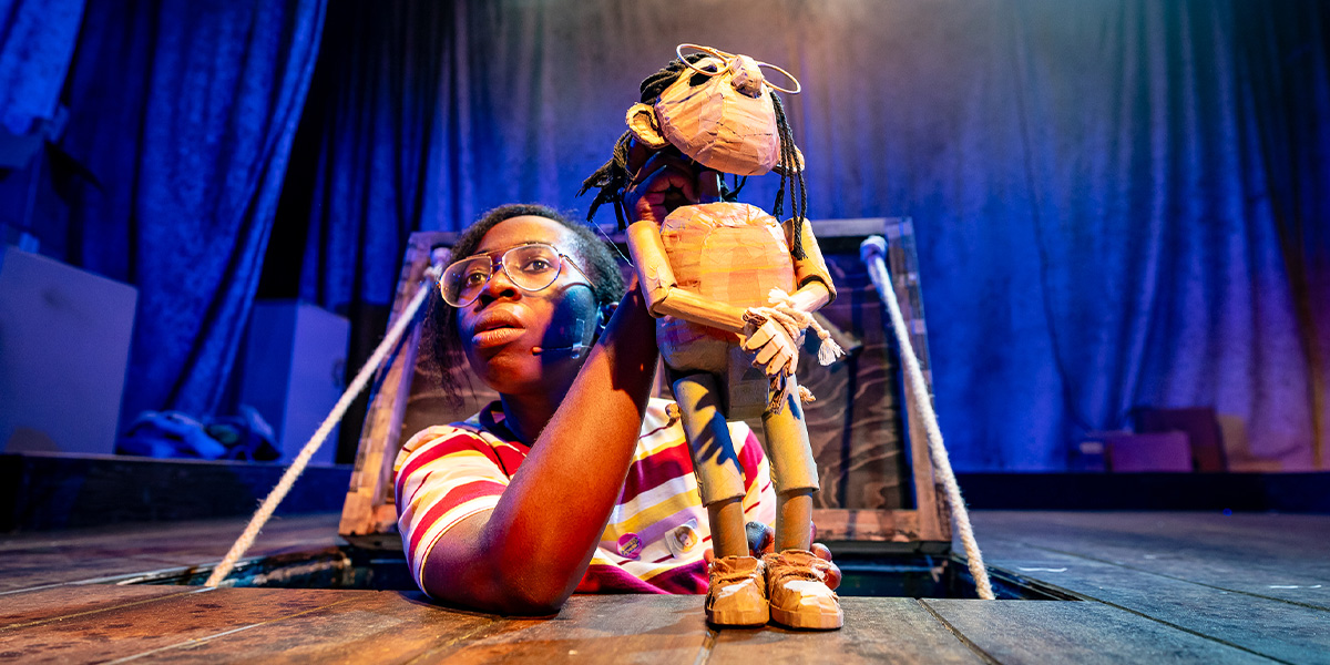 A ballet dancer in a pink tutu performs an arabesque on stage, with festive decorations in the background.