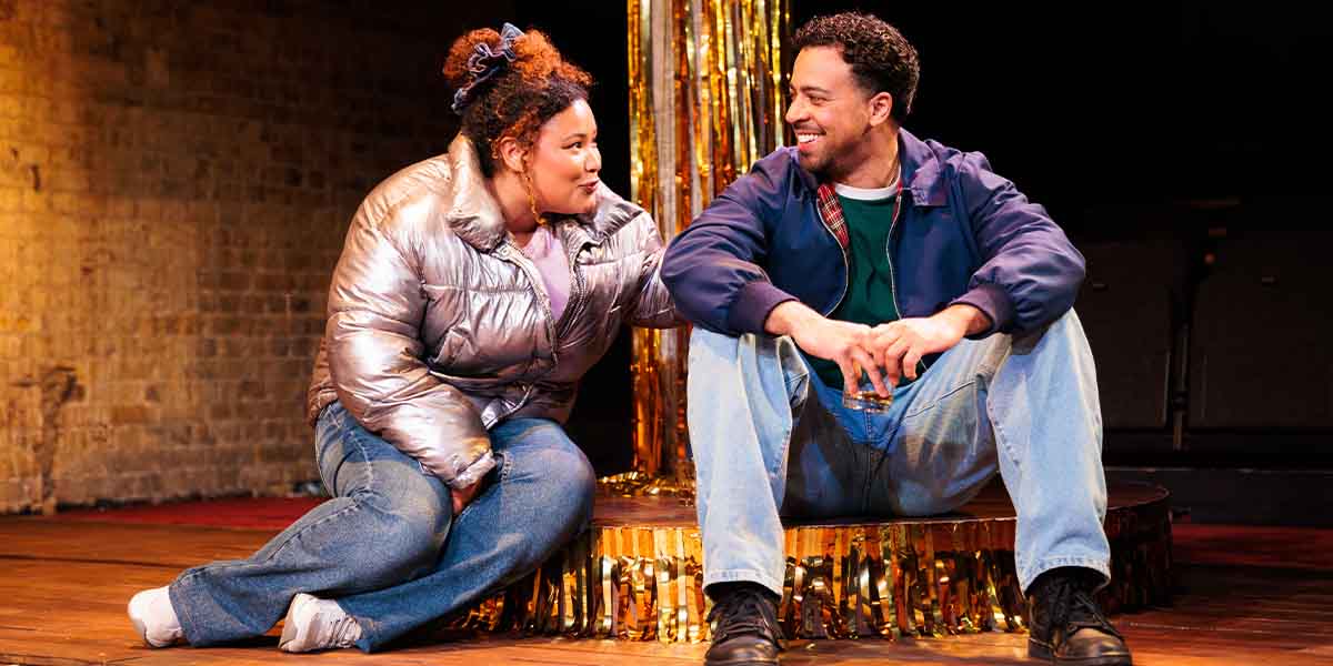 Olivia Forrest as Charlene and Aaron Anthony as Valentine sit together, smiling on stage with golden decorations.