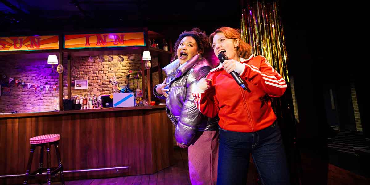 Olivia Forrest and Rowan Robinson perform energetically on stage, wearing vibrant outfits, in front of a bar.
