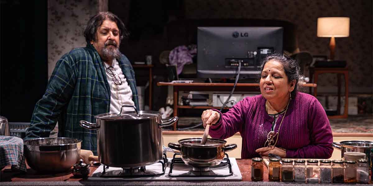 Rehan Sheikh as Liaquat watches Shobu Kapoor as Hema stirring a pot in a kitchen setting.