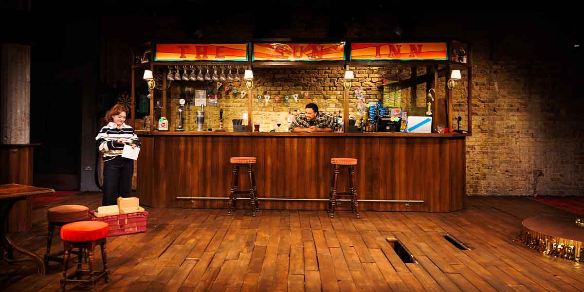 Rowan Robinson and Aaron Anthony perform at a bar set, featuring wooden stools and a brick backdrop.