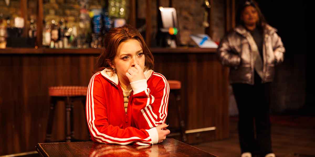 Rowan Robinson as Franky sits pensively at a table, while Olivia Forrest as Charlene stands behind.