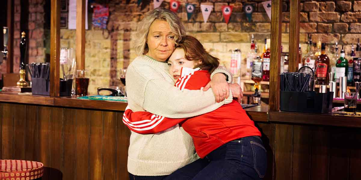 Sophie Stanton and Rowan Robinson embrace at a bar, with bottles and a brick wall in the background.