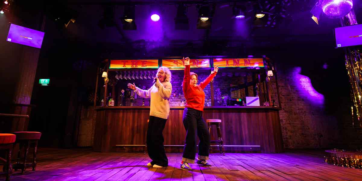 Sophie Stanton and Rowan Robinson perform energetically on stage, with a bar backdrop and vibrant lighting.
