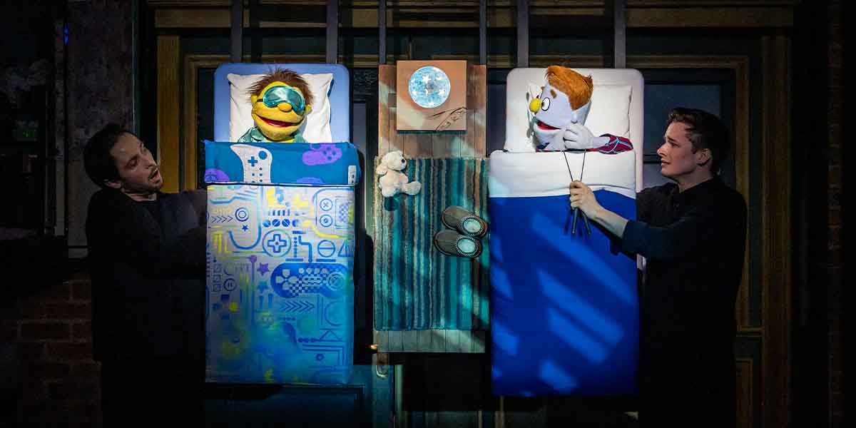 Charlie McCullagh and Noah Harrison perform with puppets in beds, featuring colourful blankets and a moonlight backdrop.