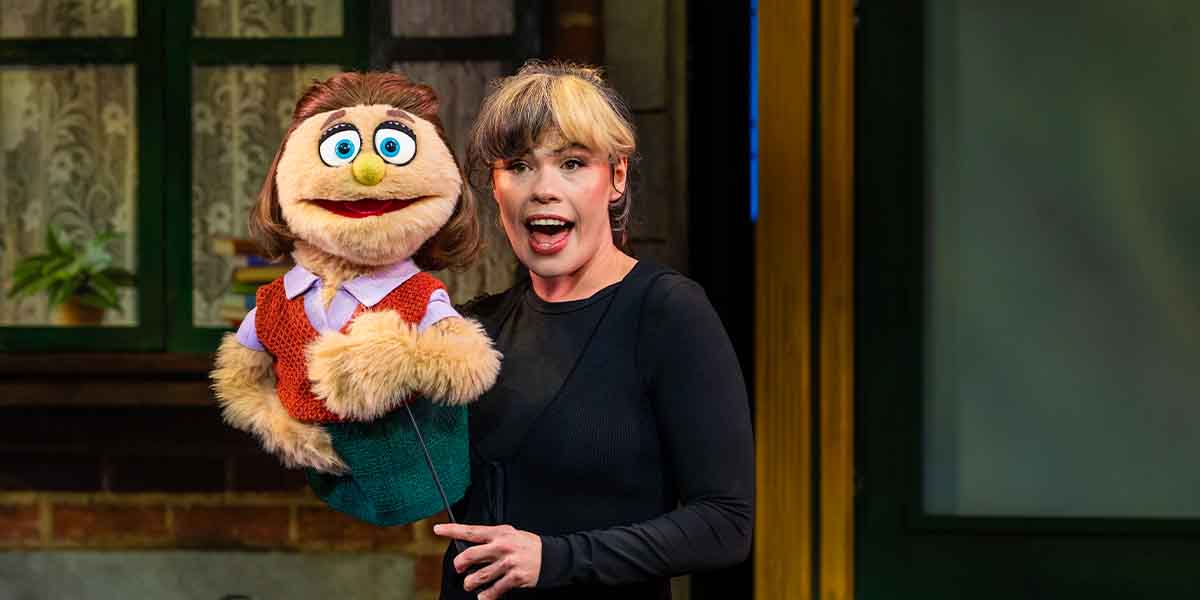 Emily Benjamin performs with a puppet, smiling and animated, against a backdrop of a stage setting.