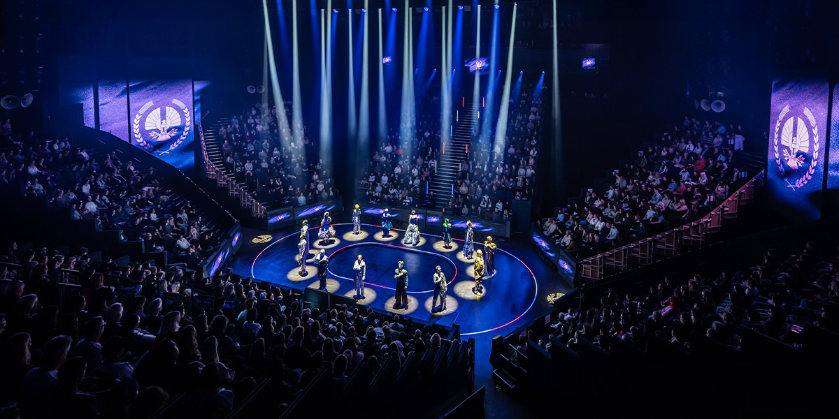 A dramatic stage scene from "The Hunger Games" featuring actors in costumes, with vibrant lighting and set design.