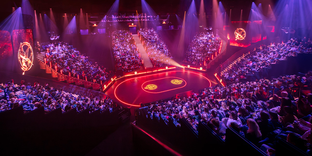 A dramatic stage scene from The Hunger Games featuring vibrant costumes, dynamic poses, and elaborate set design.