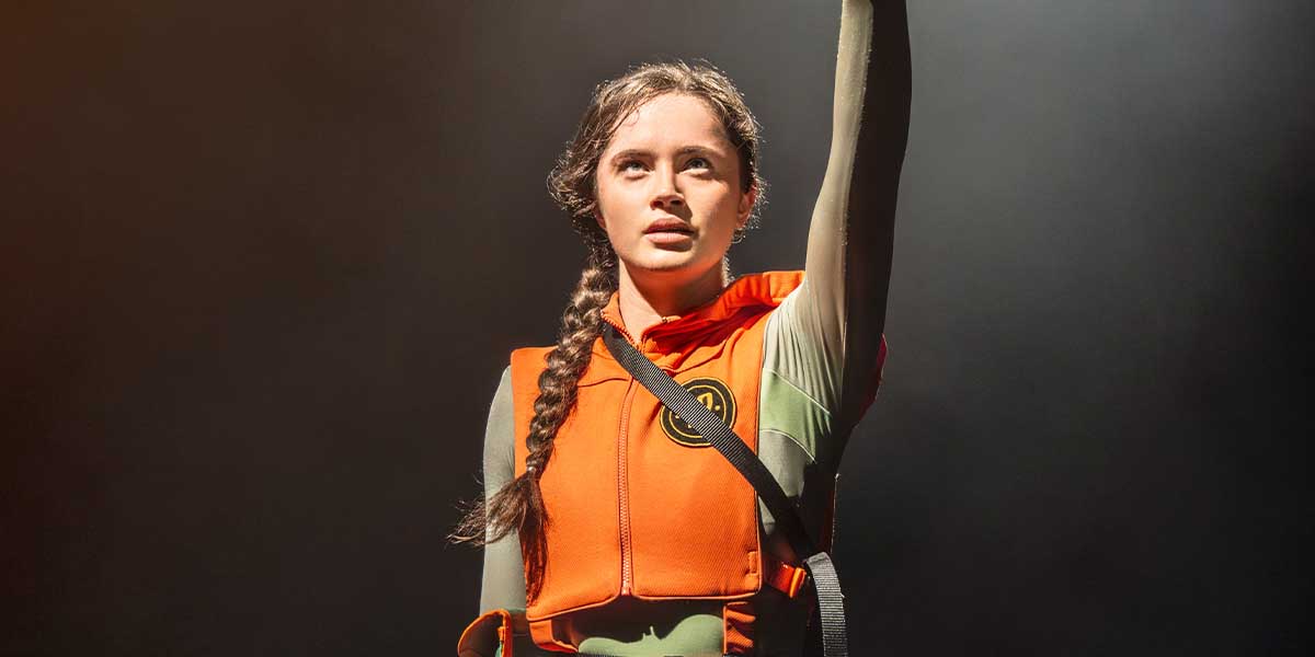 Mia Carragher as Katniss Everdeen raises her arm, wearing an orange vest against a dark background.