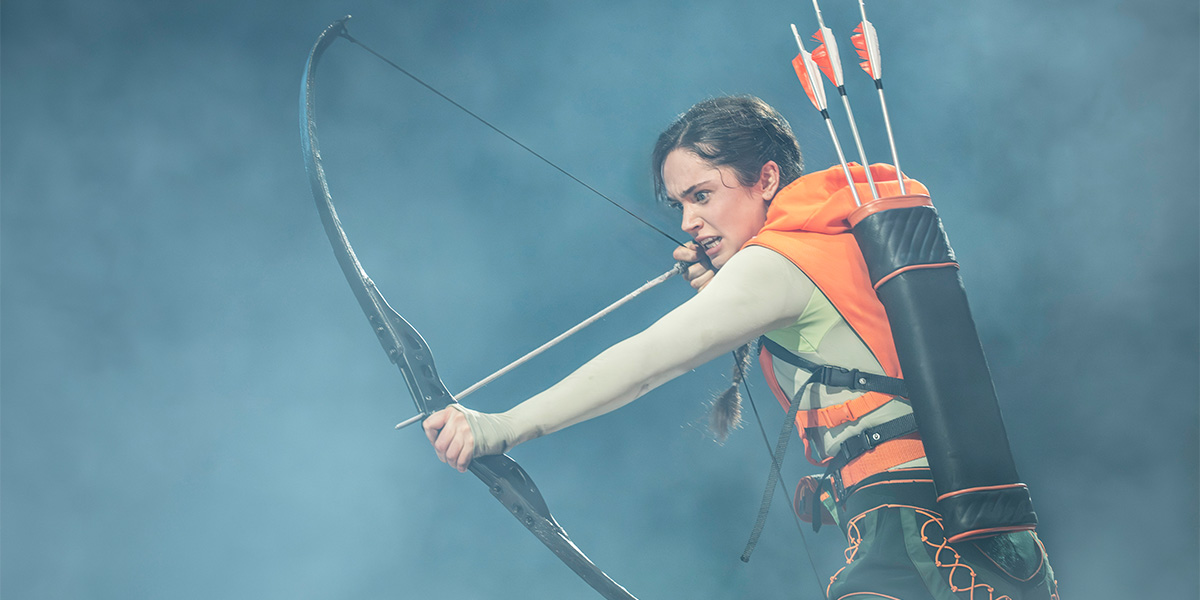 Mia Carragher as Katniss Everdeen draws a bow, wearing an orange hooded top and a quiver.