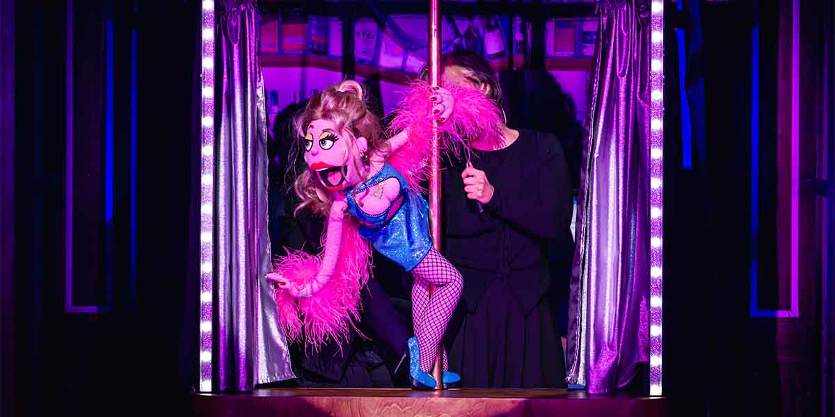 Lucy the Slut puppet, dressed in blue with pink feathers, poses on a pole, held by a performer.