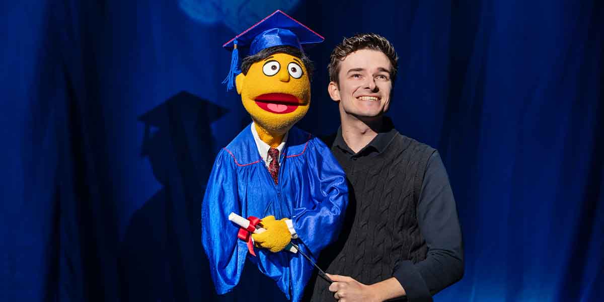 Noah Harrison smiles while holding a puppet in a graduation gown against a blue backdrop.