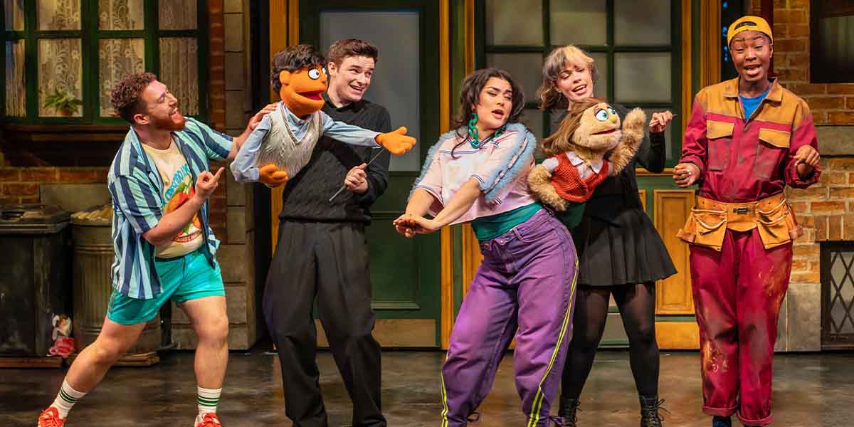 The cast of Avenue Q performs joyfully, showcasing puppets, vibrant costumes, and a lively stage backdrop.