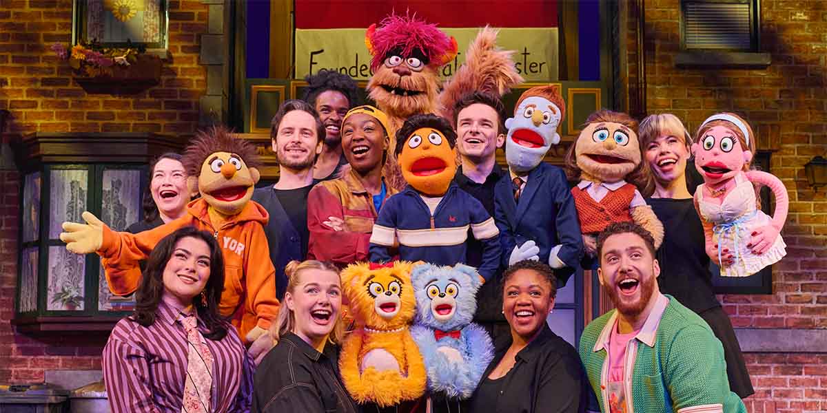The cast of Avenue Q poses together, wearing colourful costumes, with a vibrant stage backdrop.