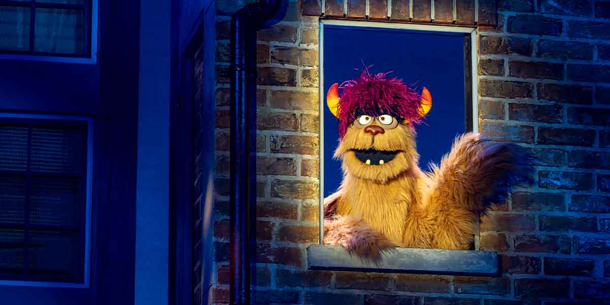 Trekkie Monster, a furry puppet with purple hair, waves from a window against a brick background.