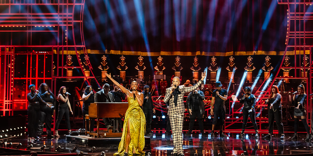 Beverley Knight and Billy Porter hosting the Olivier Awards 2025. They are standing on stage at the Royal Albert Hall. Beverley wears a yellow print dress and Billy wears a brown patterned suit.