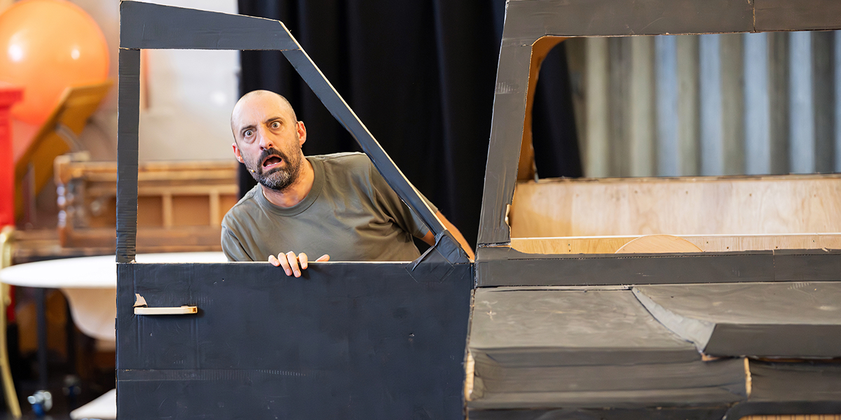 Tom Edden, portraying Mr Curry, smiles warmly while standing against a textured backdrop in a theatrical setting.