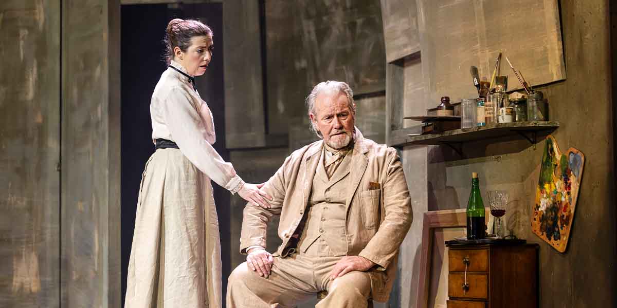 Natalie Day stands beside Jeff Shankley, who sits pensively, in a theatrical setting with muted colours.