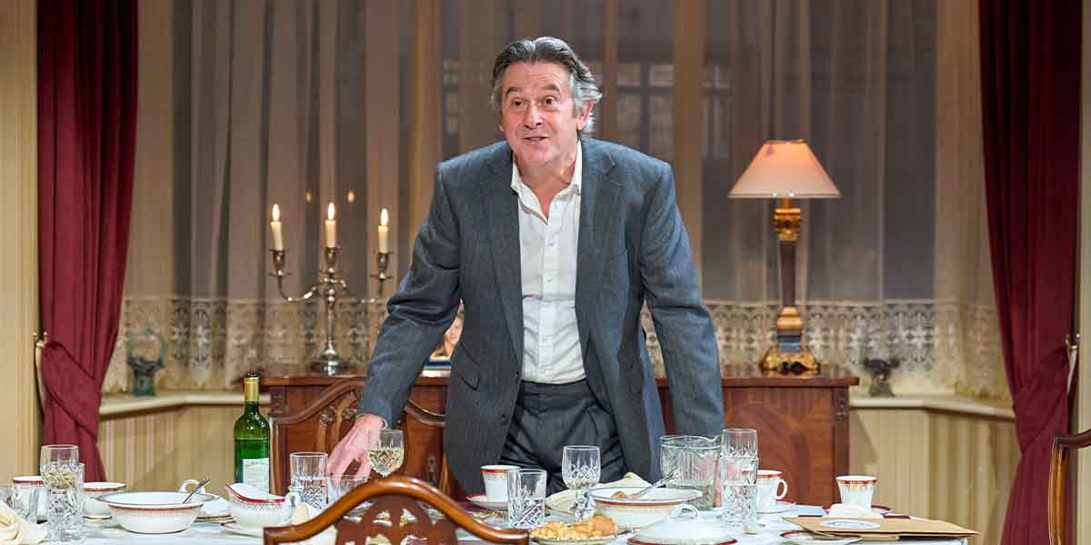 Adrian Lukis, as Sir Stephen Crossley, stands at a dining table set with glassware and food.