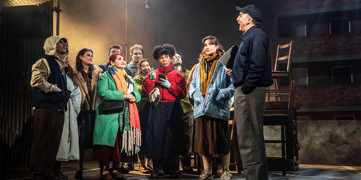 A group of actors in colourful costumes gathers around a man speaking passionately on stage.