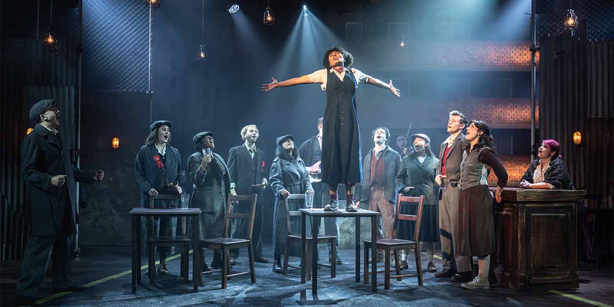 A performer stands on a table, arms outstretched, surrounded by a singing ensemble in dramatic lighting.