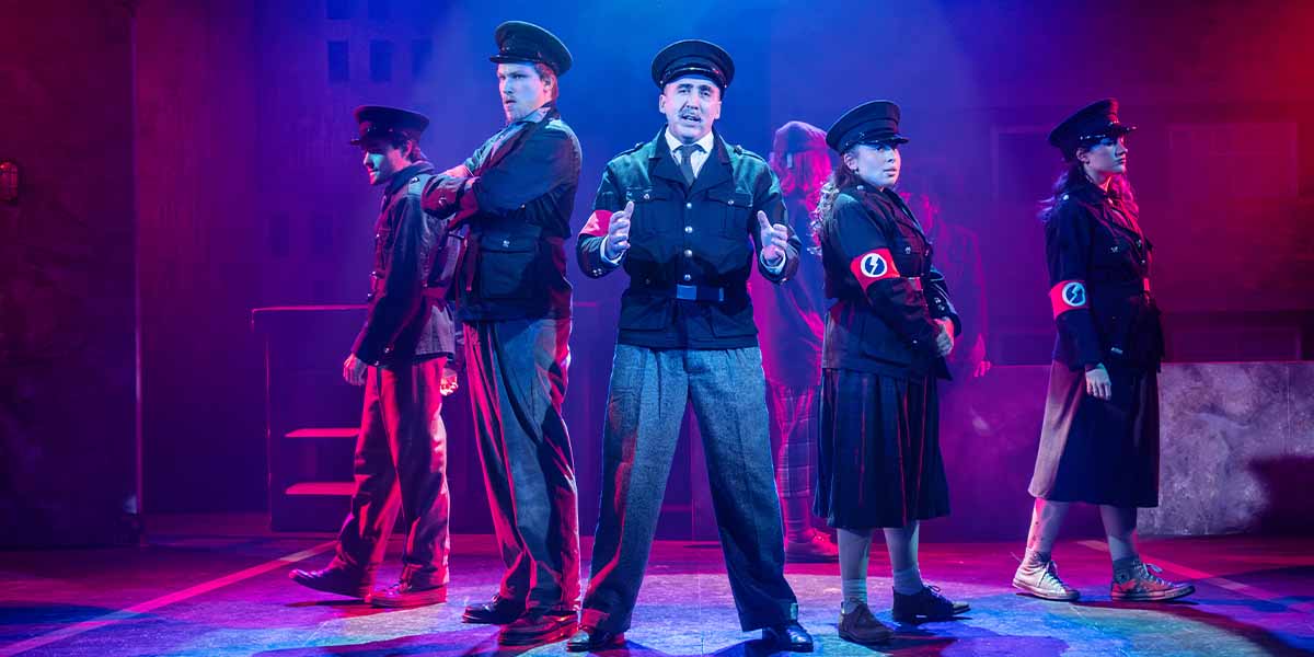 Six performers in military-style uniforms stand in a dramatic pose against a colourful, smoky backdrop.