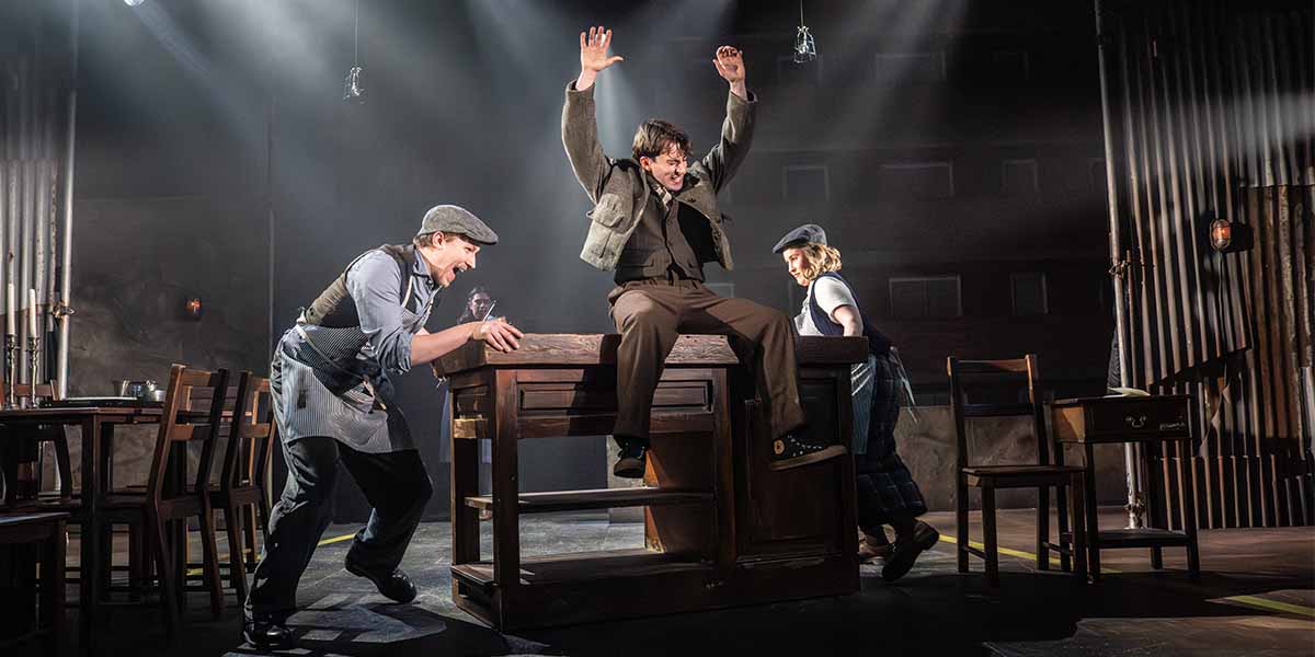 Three actors interact dramatically around a wooden table, with one jumping up and raising arms.