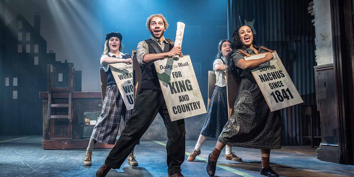Four performers in period costumes hold newspapers, smiling and singing against a theatrical backdrop.