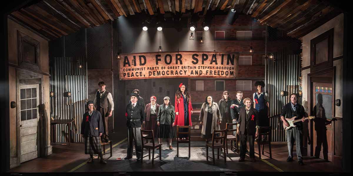 A group of actors stands on stage, with a banner reading "Aid for Spain" in the background.