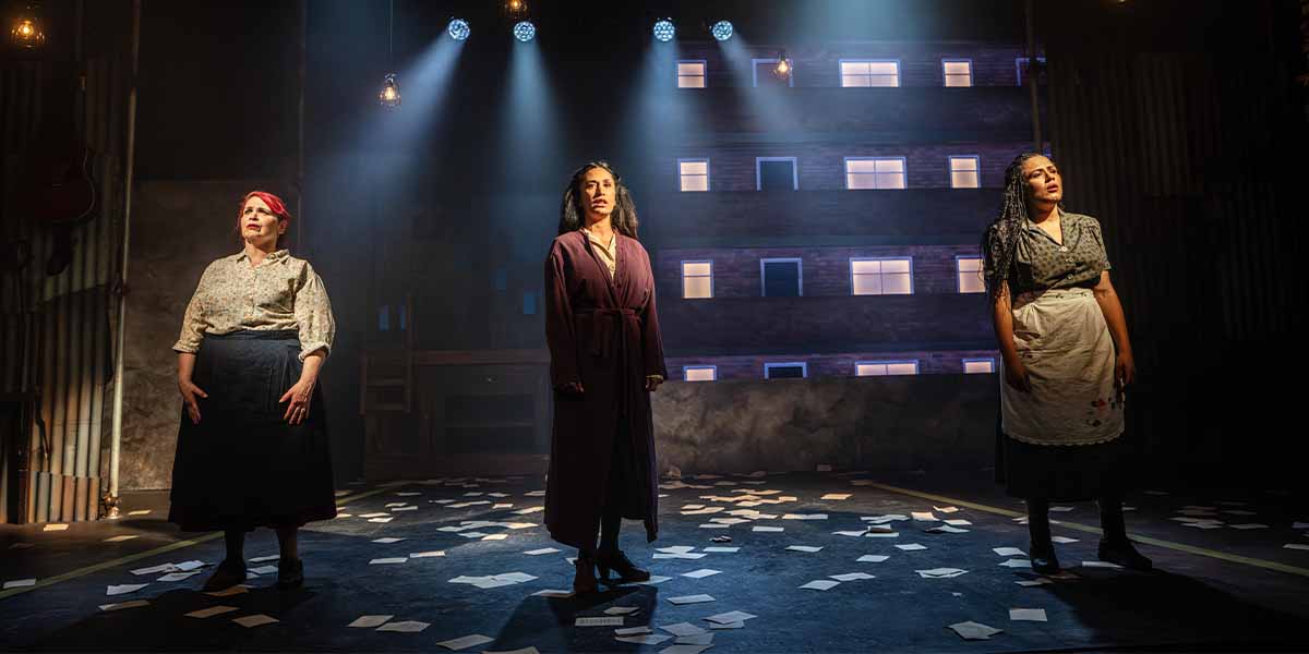 Three women stand on stage, illuminated by spotlights, surrounded by scattered papers and a dark backdrop.
