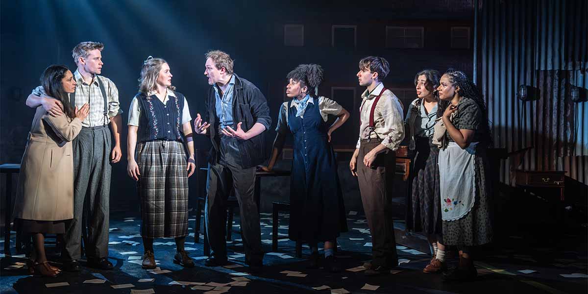A group of eight actors in period costumes, engaged in an intense discussion on a dimly lit stage.