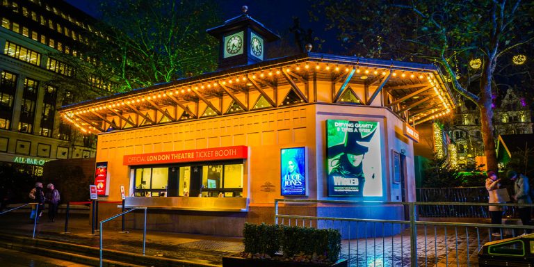 Illuminated ticket booth at night, featuring vibrant posters and a clock tower against a dark sky.