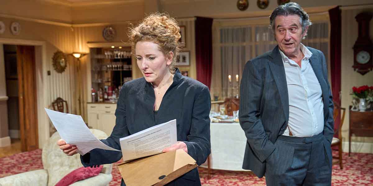 Dorothea Myer-Bennett as Ruth Rosenberg reads documents, while Adrian Lukis as Sir Stephen Crossley observes.