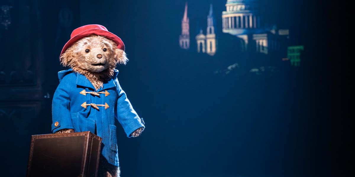 Paddington Bear, wearing a blue coat and red hat, stands with a suitcase against a dark backdrop.