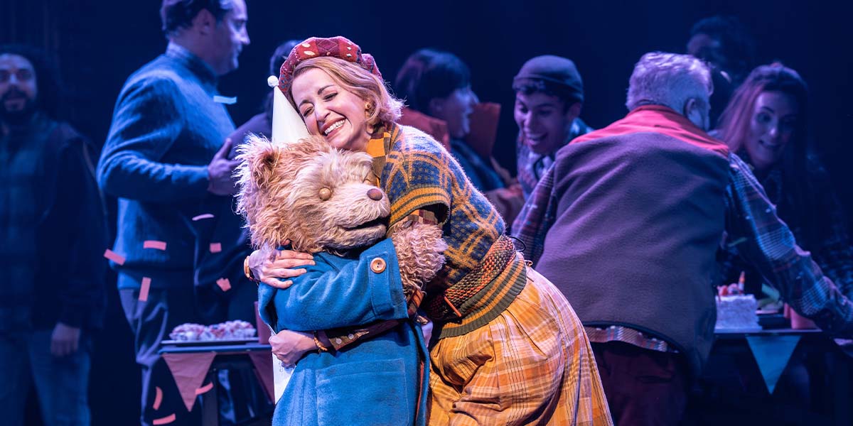 A scene from Paddington The Musical featuring Paddington Bear in a blue coat and red hat, surrounded by vibrant stage decorations.