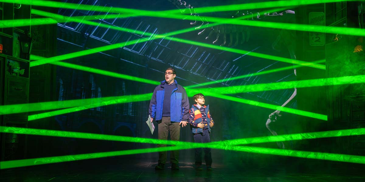 A scene from Paddington The Musical featuring Paddington Bear in a blue coat and red hat, smiling.