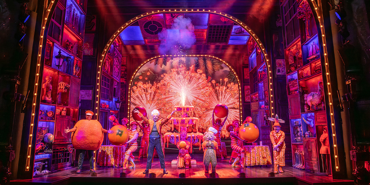 A scene from Paddington The Musical featuring Paddington Bear in a blue coat and red hat on stage. Theatre Tickets for Paddington are available from Official London Theatre.