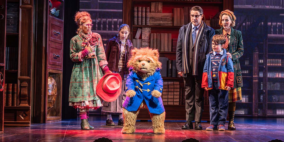 A vibrant scene from Paddington The Musical, featuring Paddington Bear in a red hat and blue coat.
