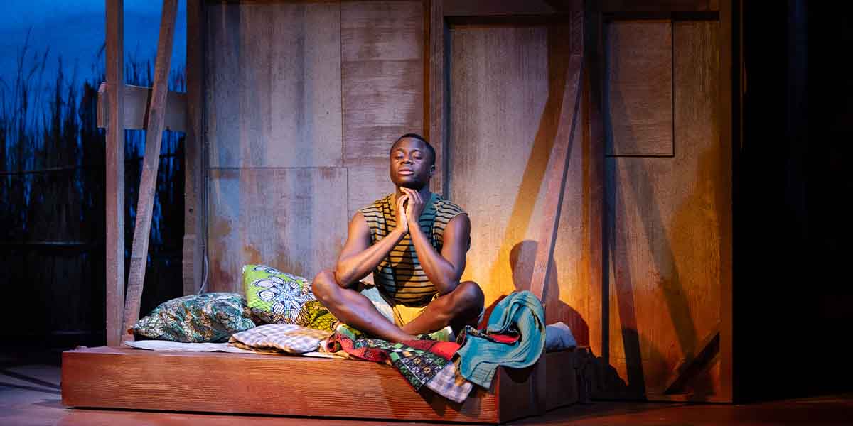 A person sits cross-legged on a wooden platform, surrounded by colourful cushions, in a dimly lit setting.