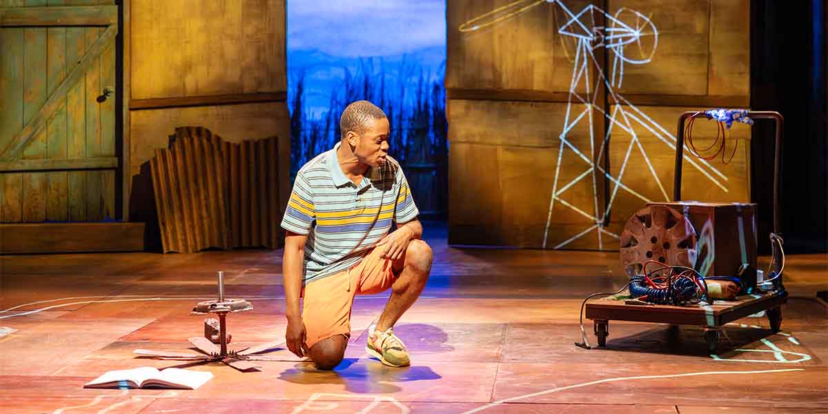 A person kneels on stage, examining a mechanical device, with a wooden backdrop and projected images.
