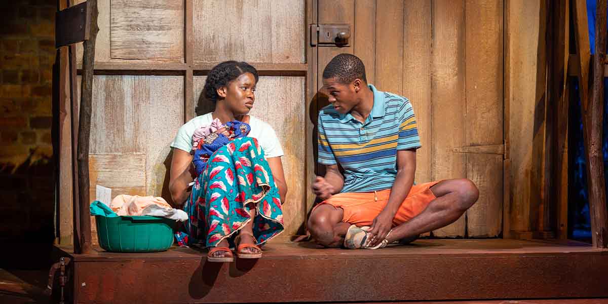 Two people sit on a wooden platform, engaged in conversation, with vibrant clothing and a rustic backdrop.